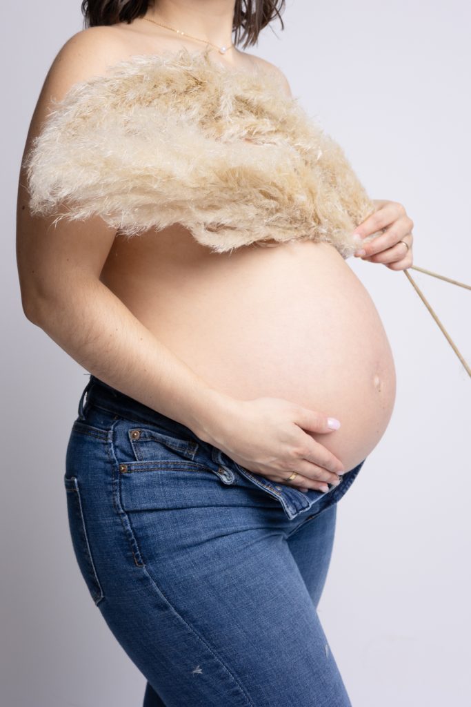 Ventre d’une femme enceinte en jeans avec des fleurs fors d’une séance en studio grossesse en Moselle.