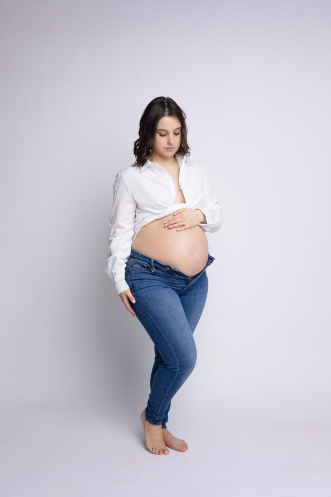 Portrait d’une femme enceinte en jeans et chemise blanche lors d’une séance grossesse en studio en Moselle.
