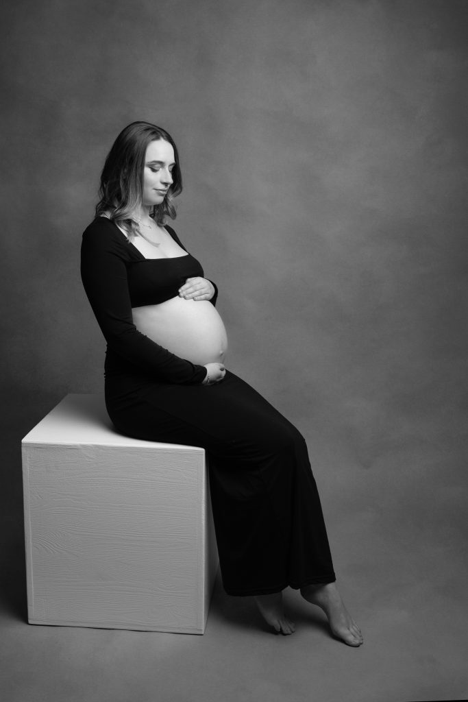 femme en séance photo qui attend un enfant et qui regarde son ventre assise sur un cube blanc