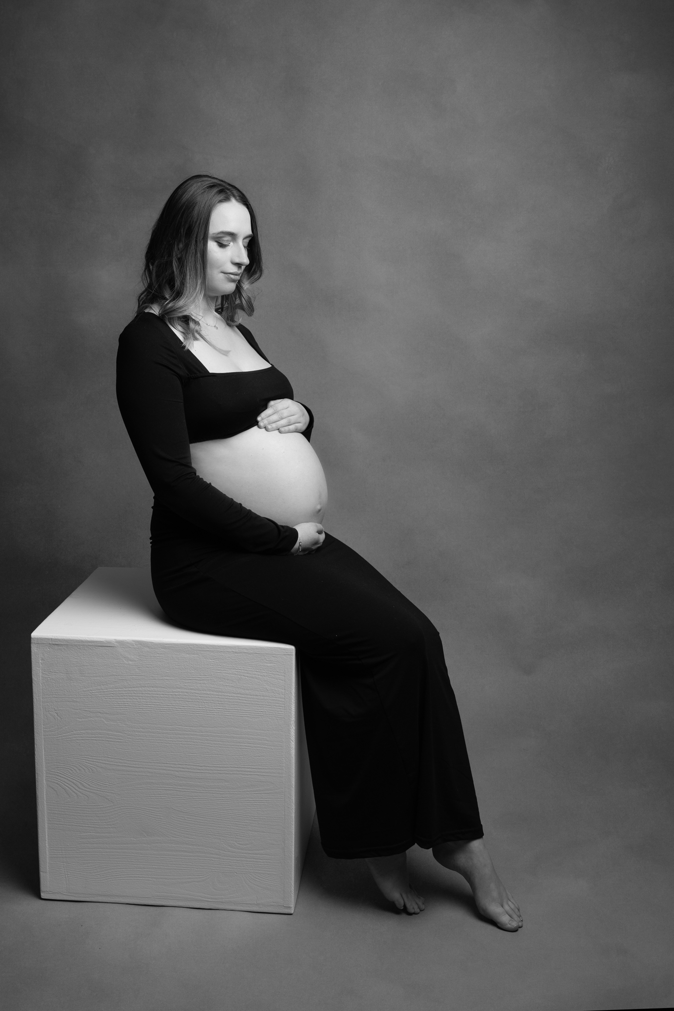 femme en séance photo qui attend un enfant et qui regarde son ventre assise sur un cube blanc